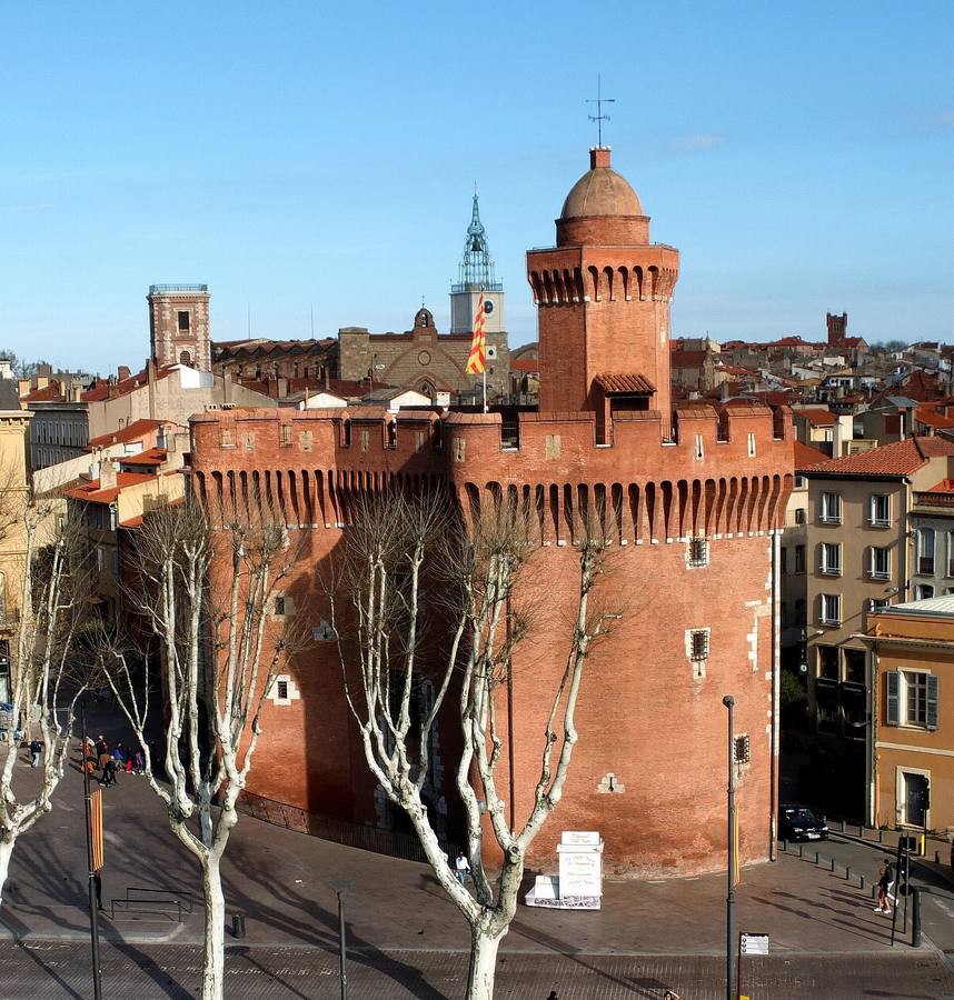 Le Castillet de Perpignan, porte fortifiée en brique rouge surmontée d'une tour à coupole, avec le clocher de la cathédrale à l'arrière-plan